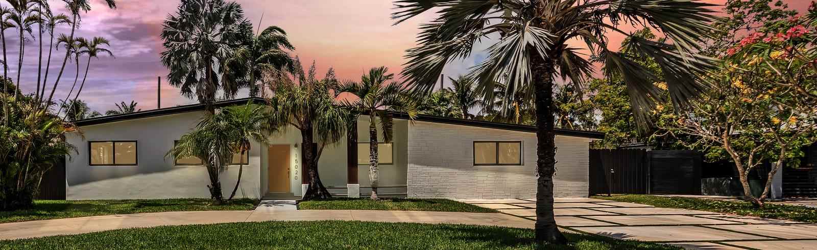 Renovated single-story South Florida home at dusk with a white stone facade, mature palm trees and a paver driveway